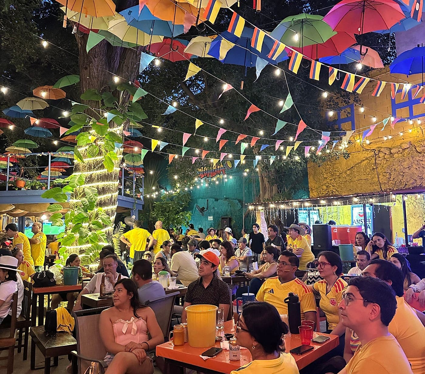 Terraza de Parceros llena de comensales una noche de partido, bajo el techo de sombrillas, los banderines y la columna de luces