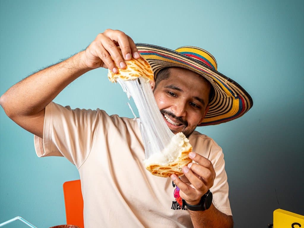 A man in a sombrero vueltiao pulls apart a fresh arepa, stretching the melted cheese between the two halves.