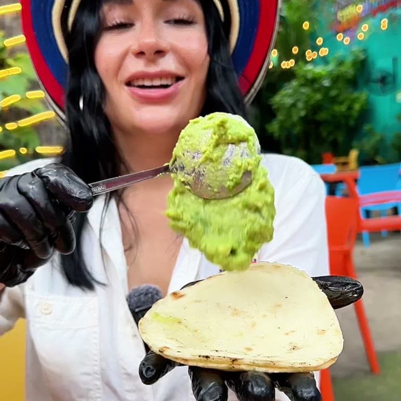 A freshly made arepa tortilla held in a hand, with a spoonful of guacamole dropping onto it.