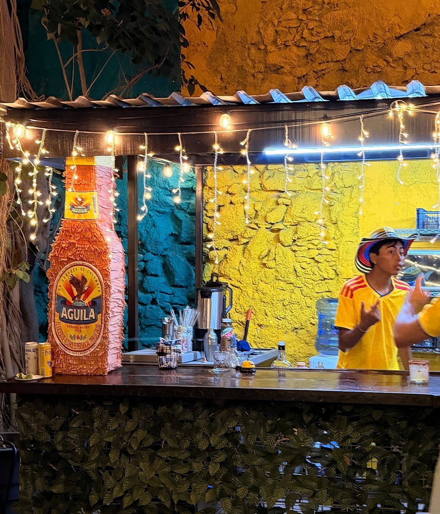 La barra del patio iluminada de noche, contra la pared pintada como una fachada colombiana