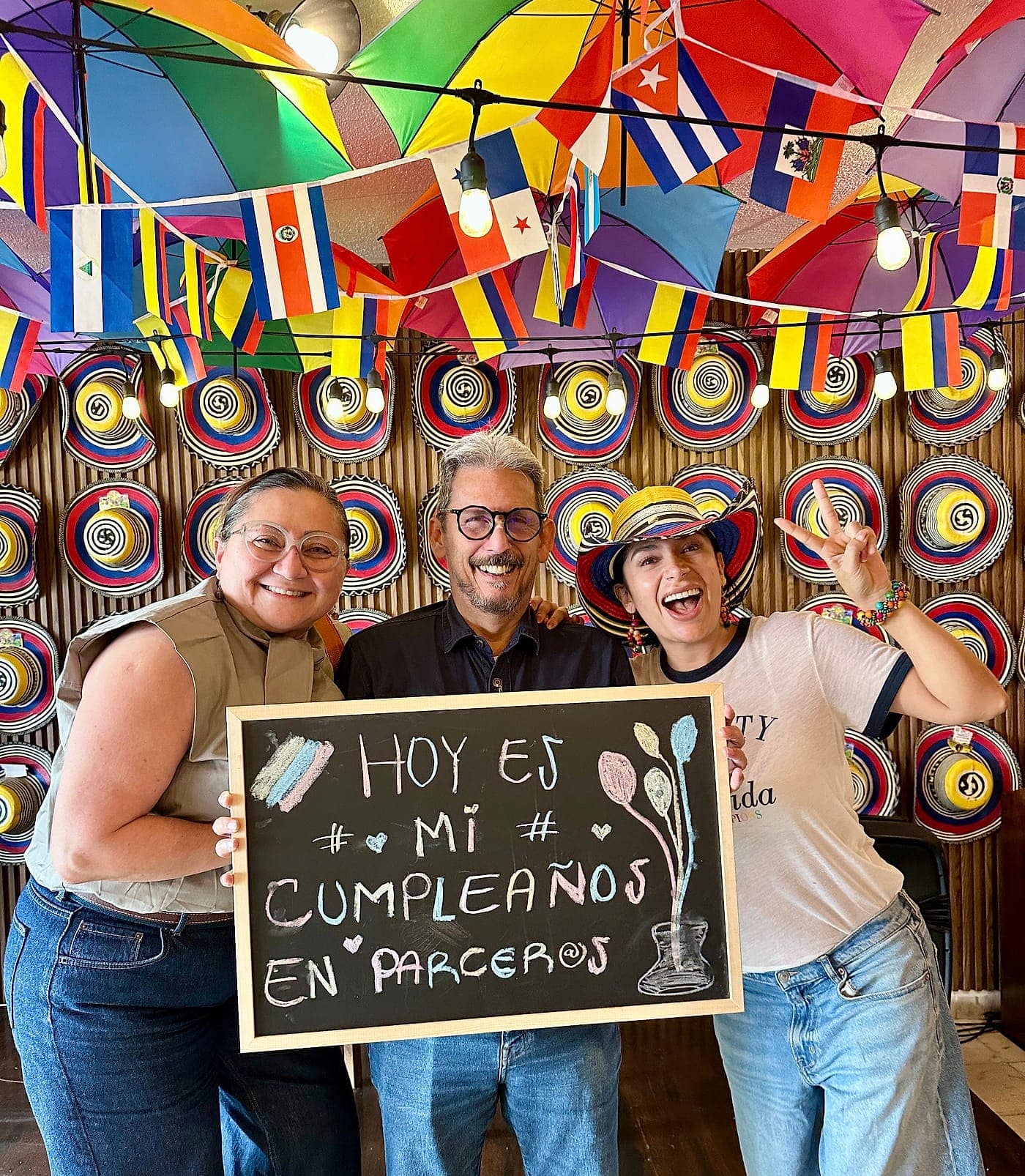 Tres personas riéndose mientras sostienen un pizarrón que dice «Hoy es mi cumpleaños en Parceros», una con sombrero vueltiao, ante la pared cubierta de sombreros y bajo las sombrillas y las banderas
