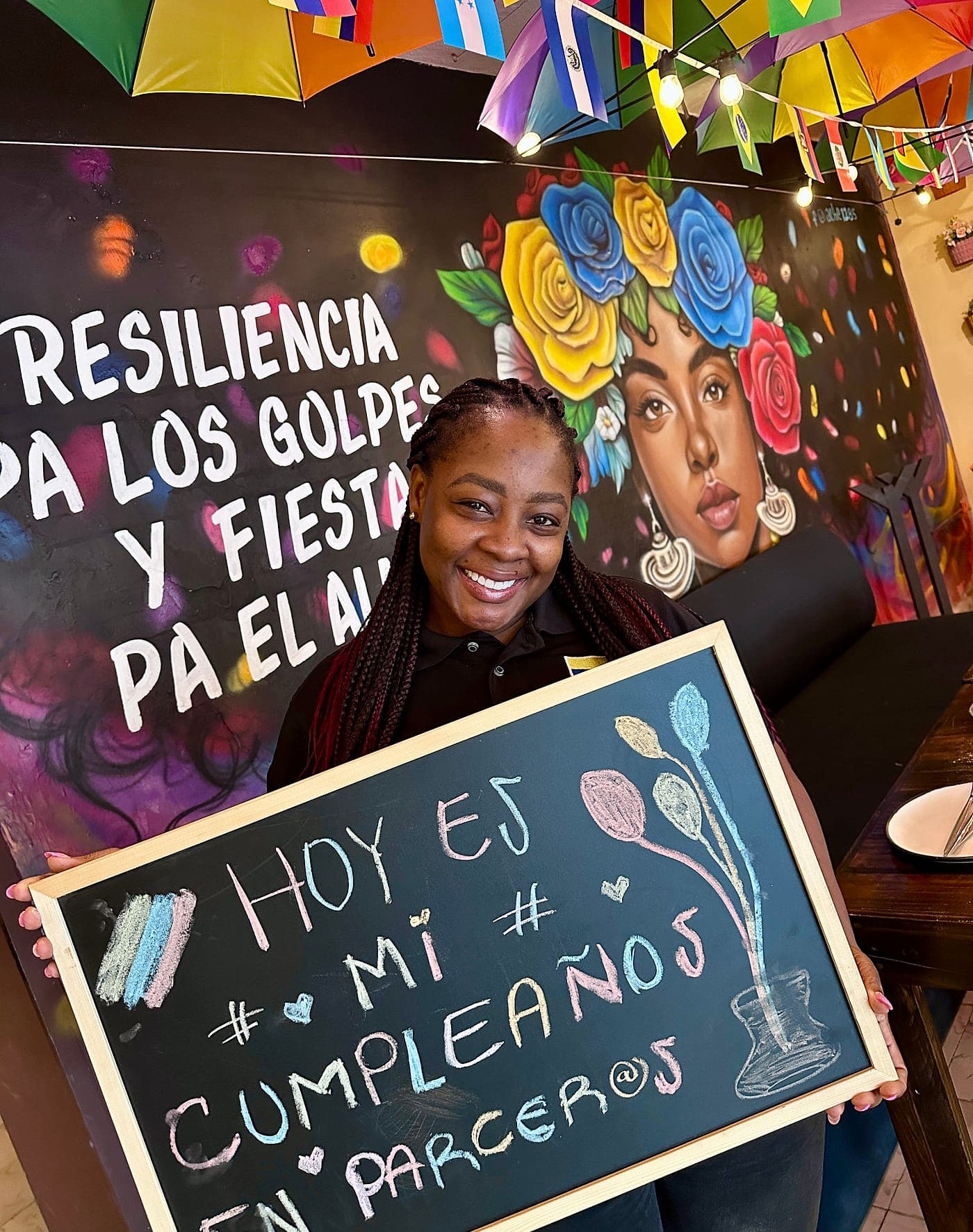 Una persona del equipo de Parceros sostiene, sonriendo, un pizarrón que dice «Hoy es mi cumpleaños en Parceros» delante del mural «Resiliencia pa los golpes y fiesta pa el alma»