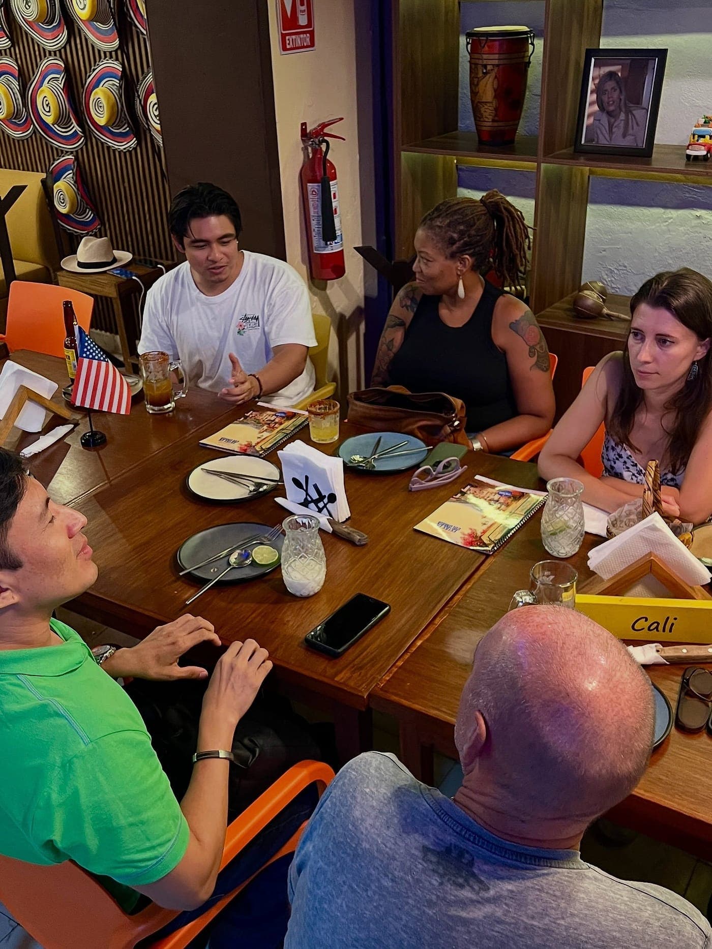 Cinco personas conversando alrededor de una mesa con cuadernos abiertos, una banderita de Estados Unidos y el letrero de mesa «Cali», ante la pared de sombreros vueltiaos.