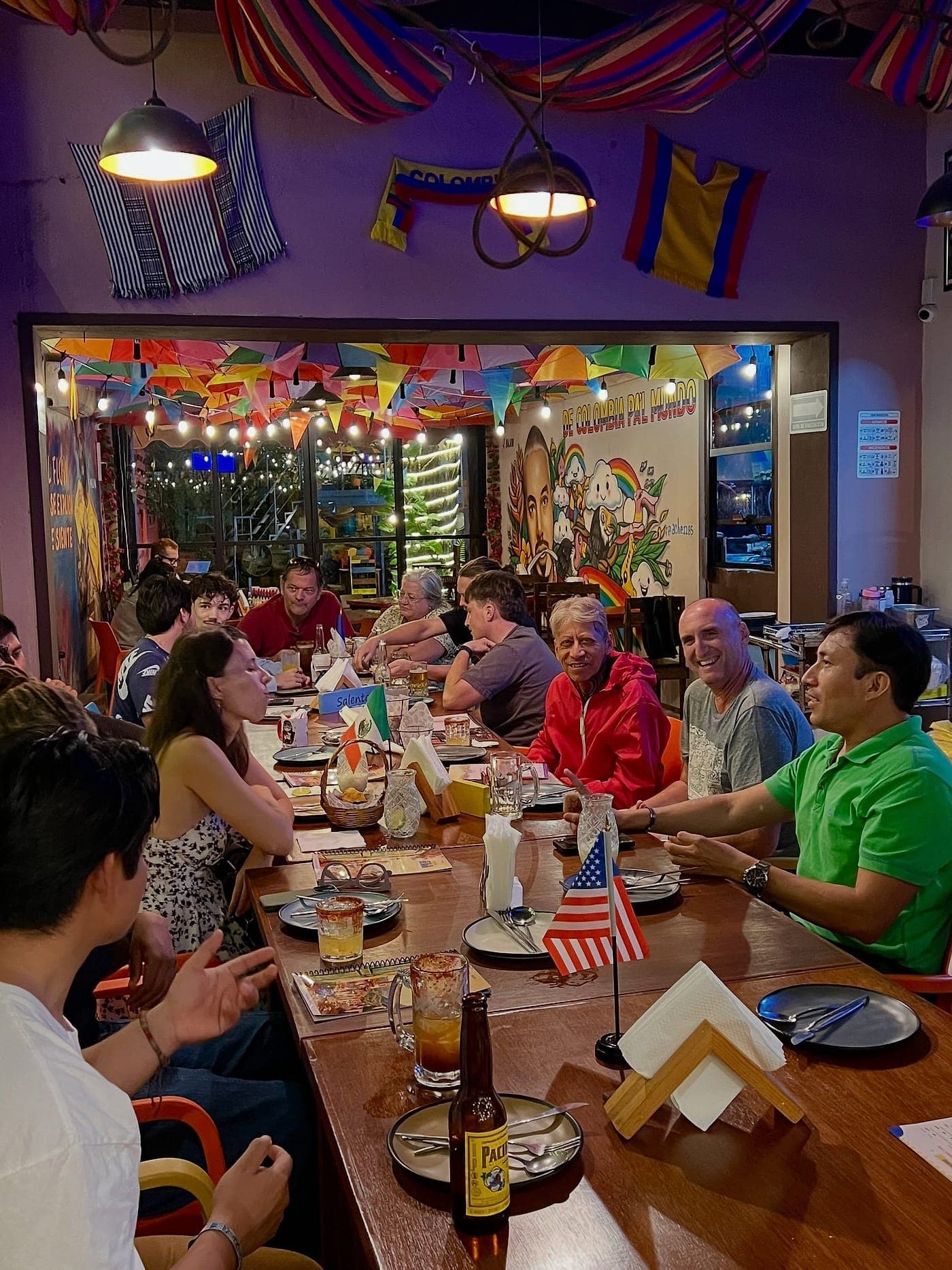 La mesa larga de Parceros llena una noche de martes: una docena de personas conversando, con banderitas de mesa de Estados Unidos e Italia y la bandera de Colombia colgada de la pared.