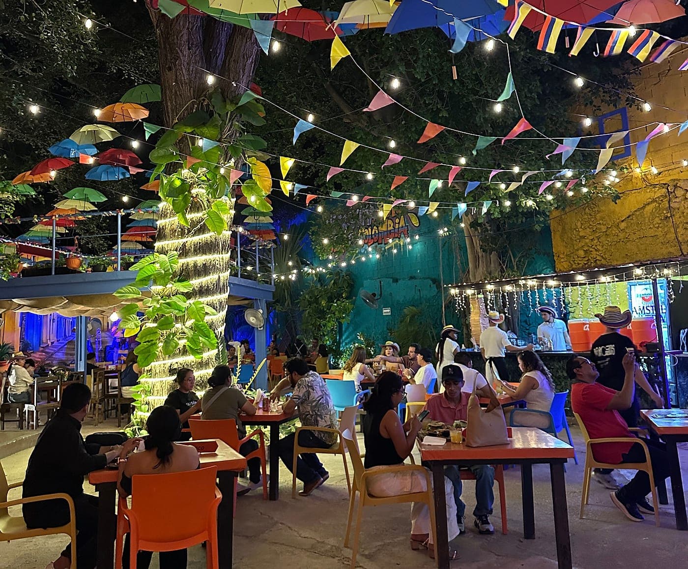 The Parceros patio at night with full tables around the column of lights, under the tree, the umbrellas and the bunting