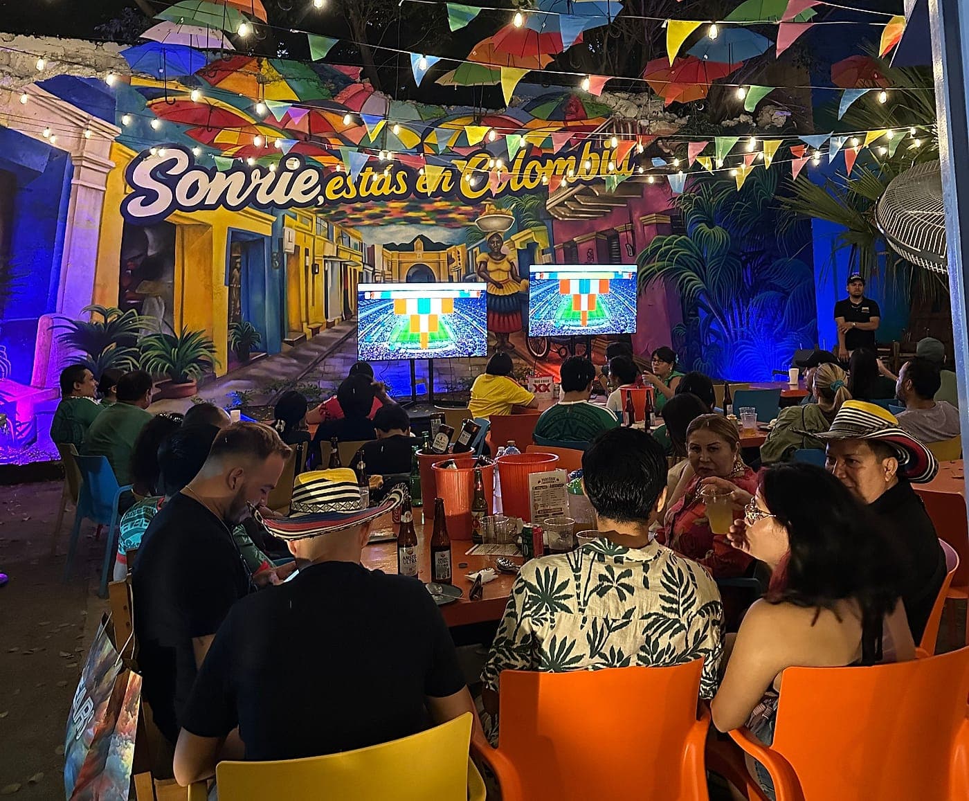 Comedor de Parceros lleno una noche de partido, con el mural «Sonríe, estás en Colombia» iluminado al fondo y el techo de sombrillas encima.