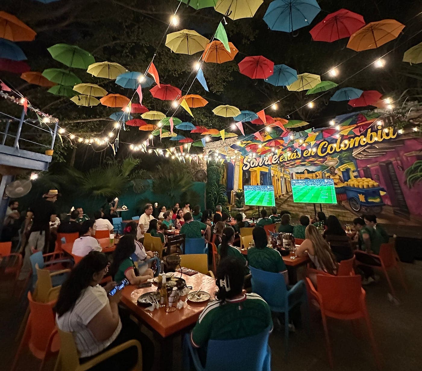 Patio de Parceros lleno de noche bajo el techo de sombrillas de colores y las guirnaldas, con el mural «Sonríe, estás en Colombia» iluminado al fondo