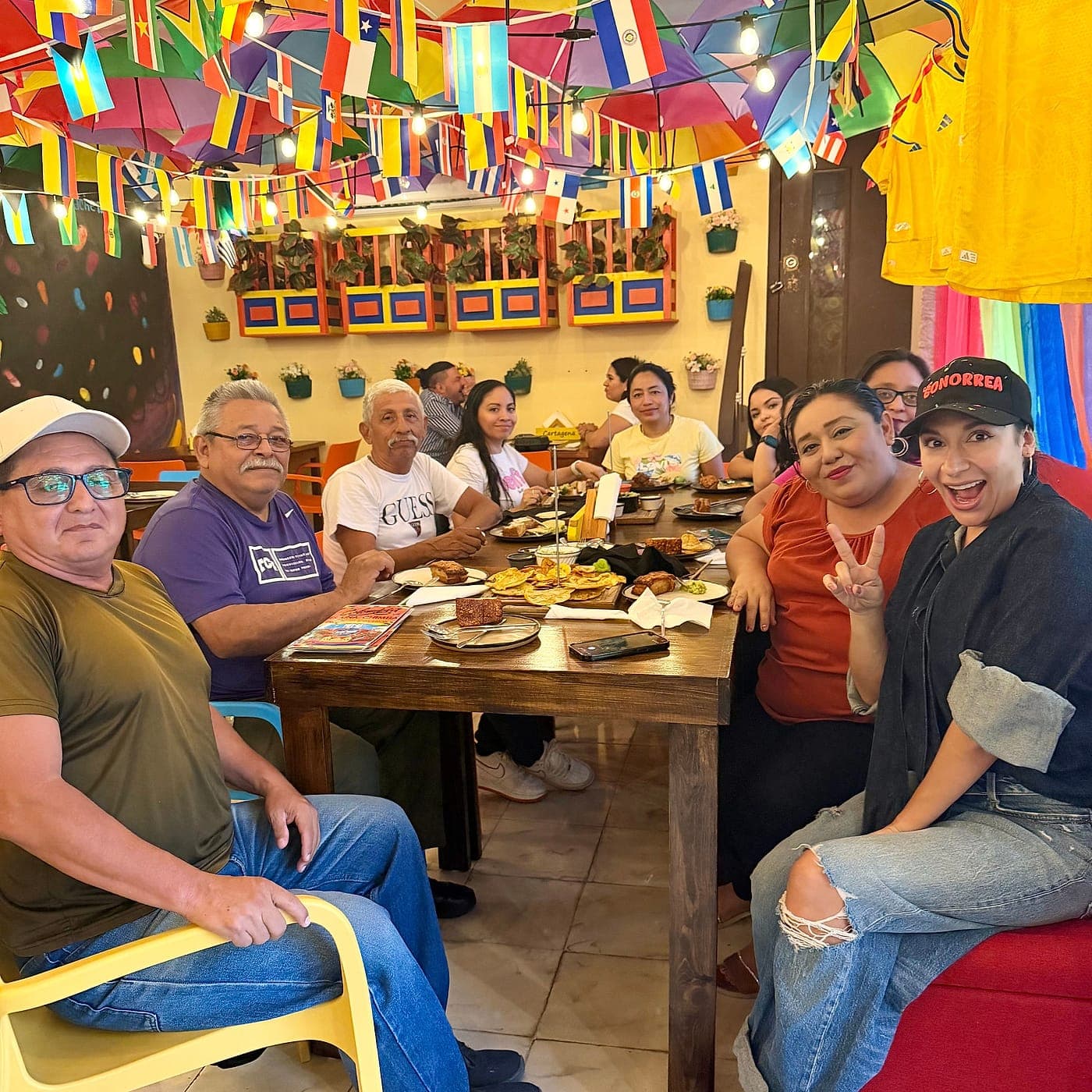Un grupo compartiendo una mesa larga de Parceros con la comida servida, bajo el techo de sombrillas y las banderas de países de América