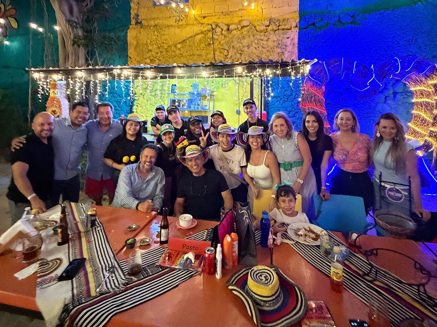 La mesa larga de Parceros llena de noche: un grupo grande posando con sombreros vueltiaos bajo el árbol con guirnaldas, ante el mural amarillo y azul, con caminos de mesa colombianos