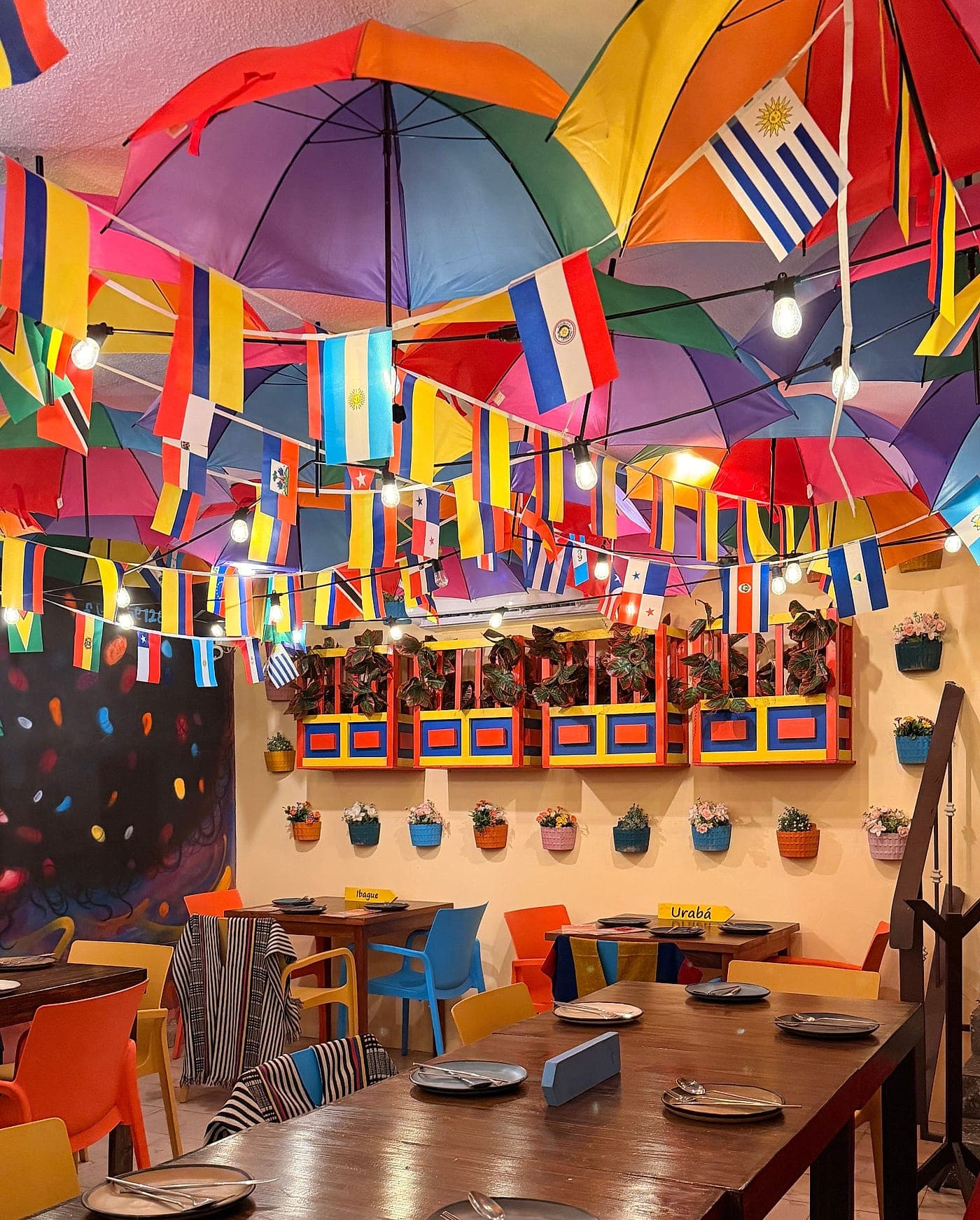 Mesa comunal de madera puesta para diez, bajo el techo interior de sombrillas de colores y banderas de países de Latinoamérica.