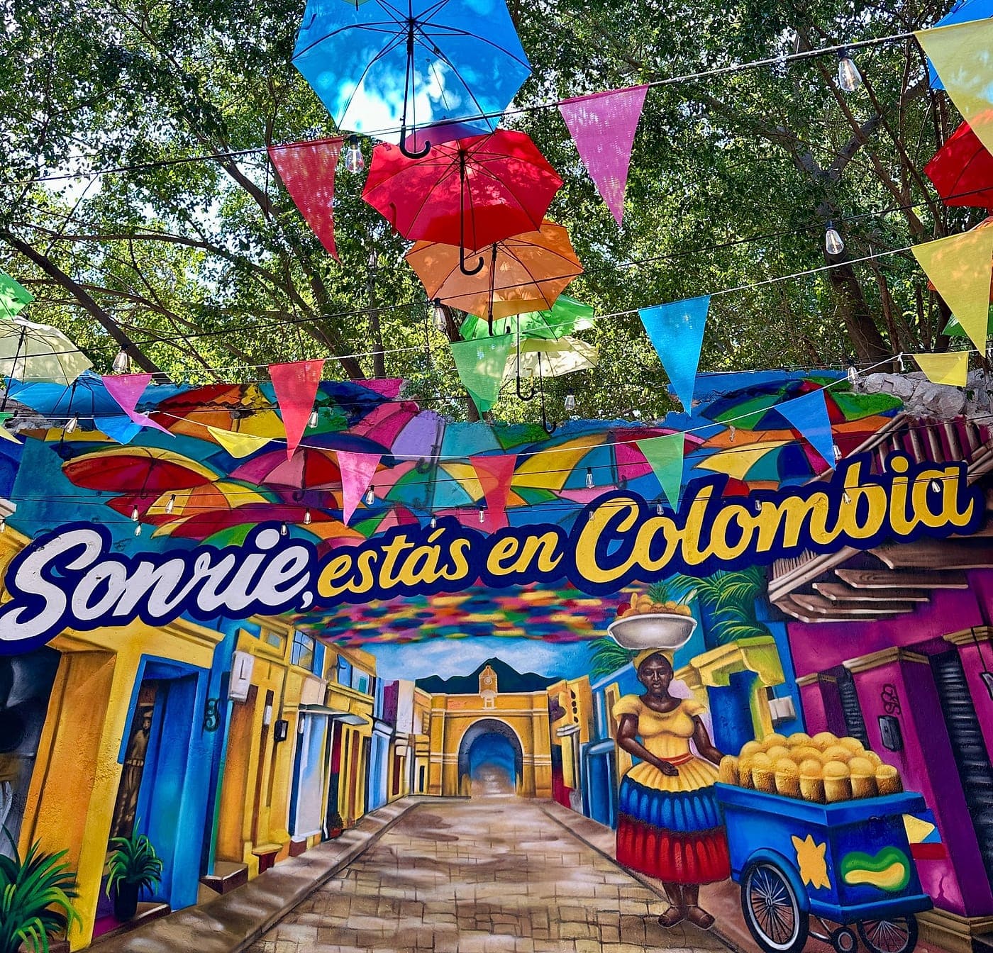 Mural de Parceros «Sonríe, estás en Colombia» a plena luz: una calle colonial pintada de colores con una palenquera y su carreta de mangos.