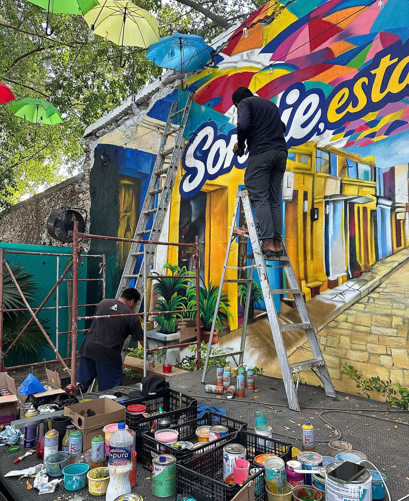 Dos muralistas subidos a una escalera pintando el mural «Sonríe, estás en Colombia», con el suelo todavía lleno de botes de pintura