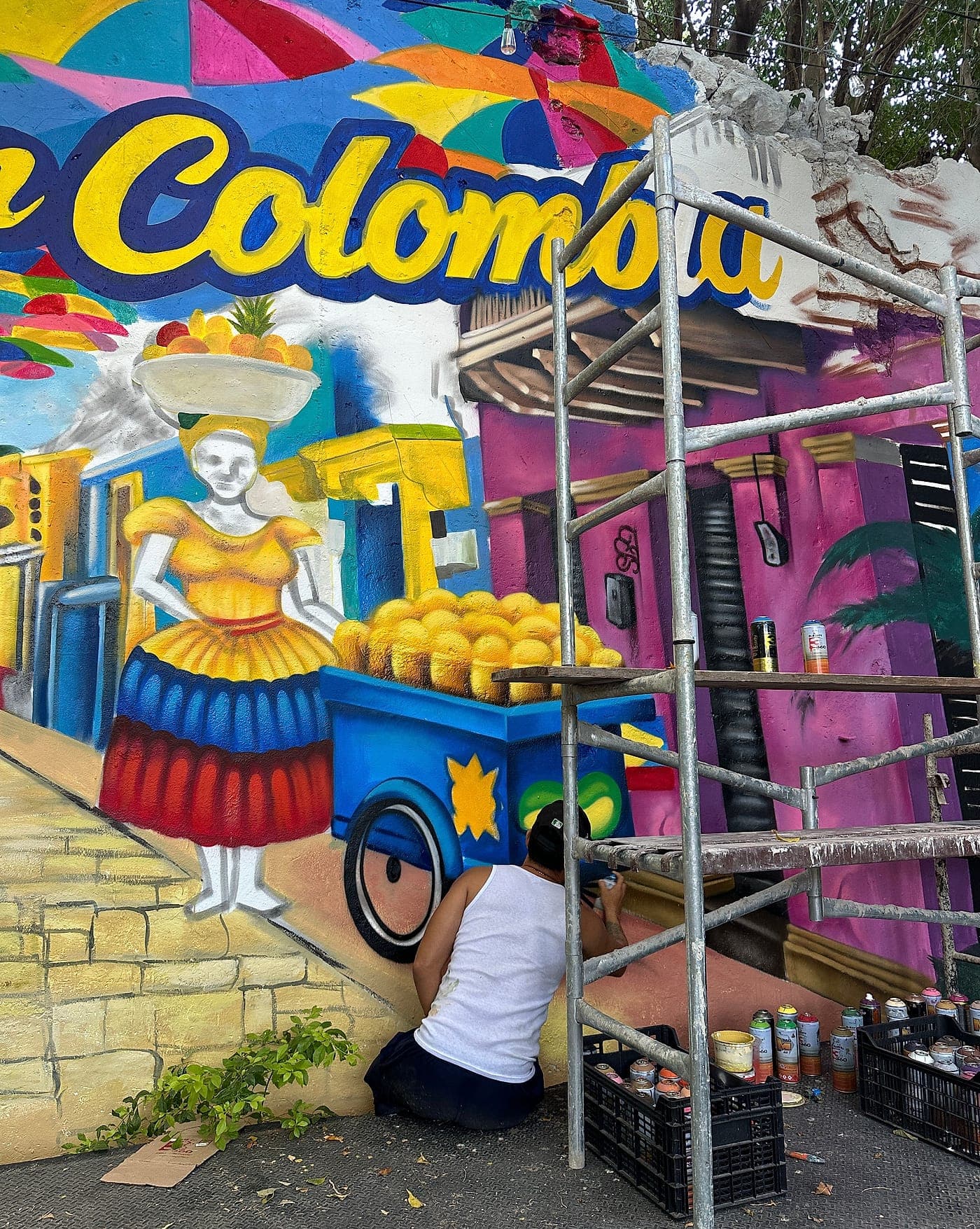 Un muralista agachado pintando la carreta de mangos de la palenquera, con el andamio montado a su lado