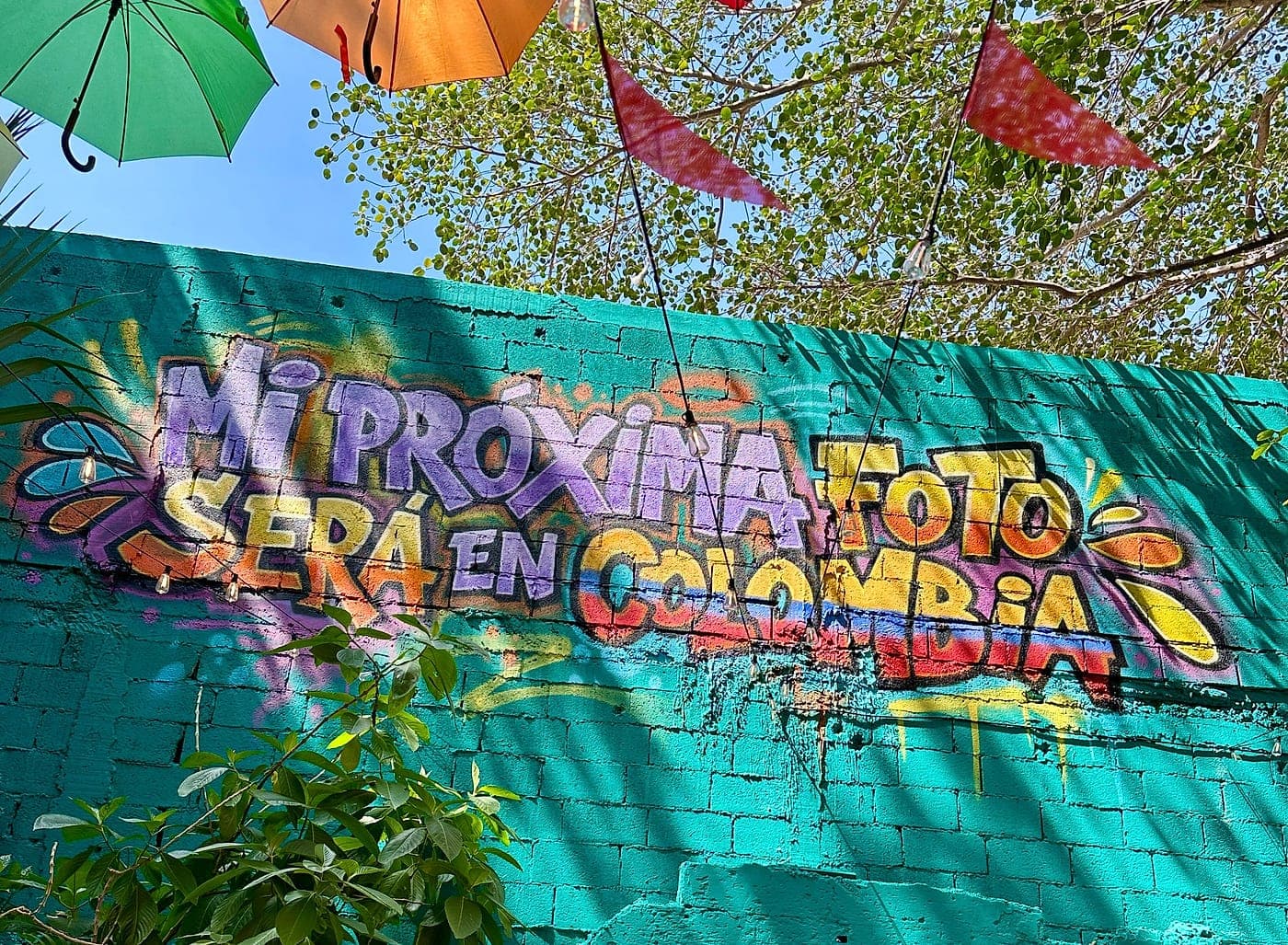Pared turquesa con el grafiti «Mi próxima foto será en Colombia», entre plantas y bajo las sombrillas de colores.
