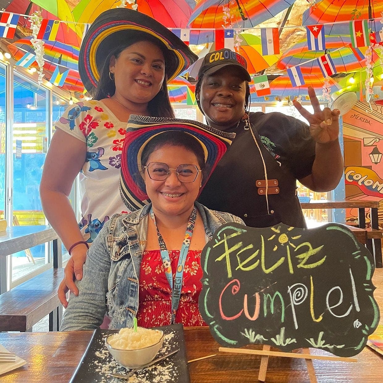 Tres personas sonriendo alrededor de un pizarrón que dice «Feliz Cumple» junto a un postre con su velita, bajo el techo de sombrillas de colores y las banderas de países