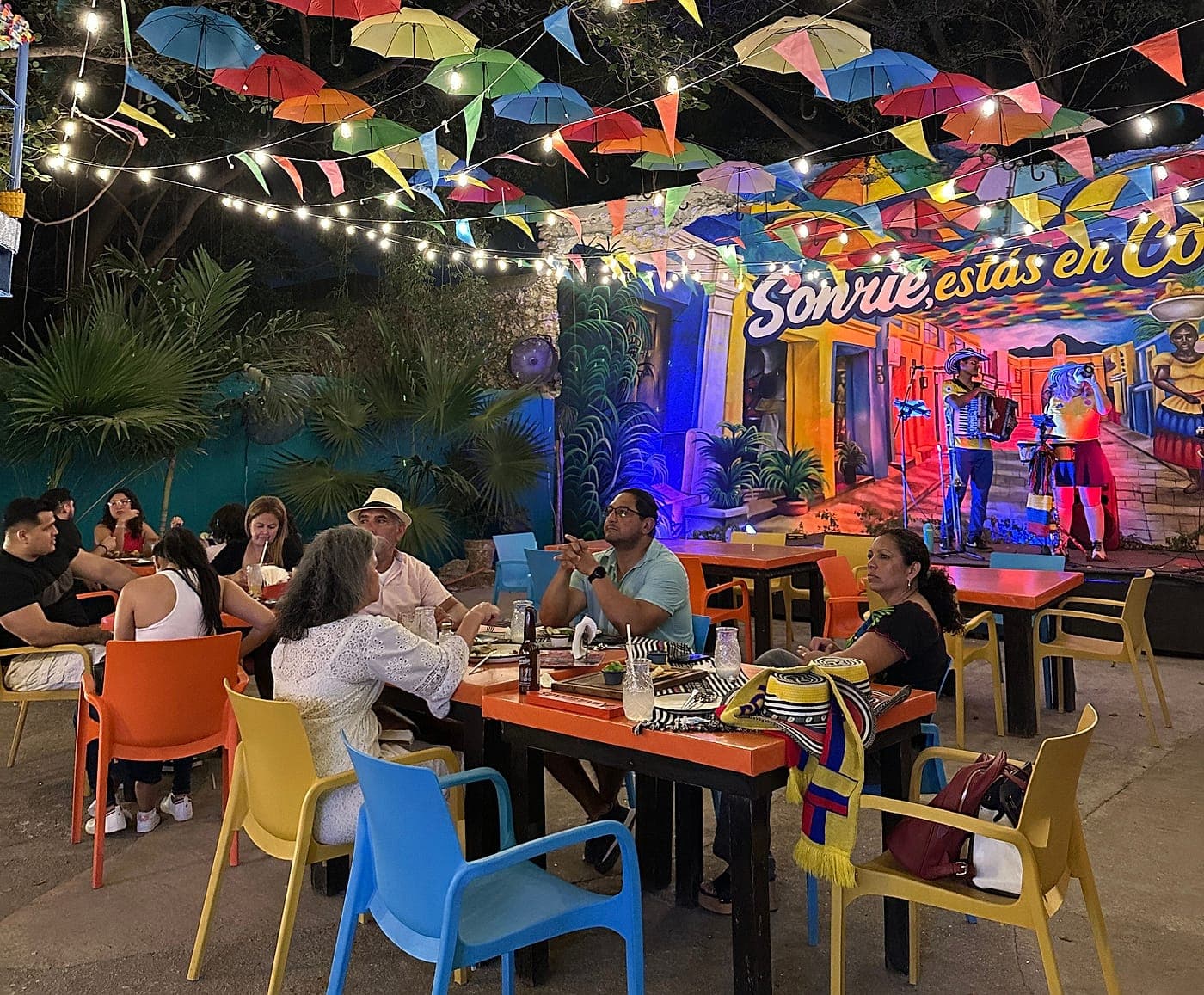 The terrace at Parceros at night with the tables full, the mural lit up and a musician playing at the back, under the umbrella ceiling.