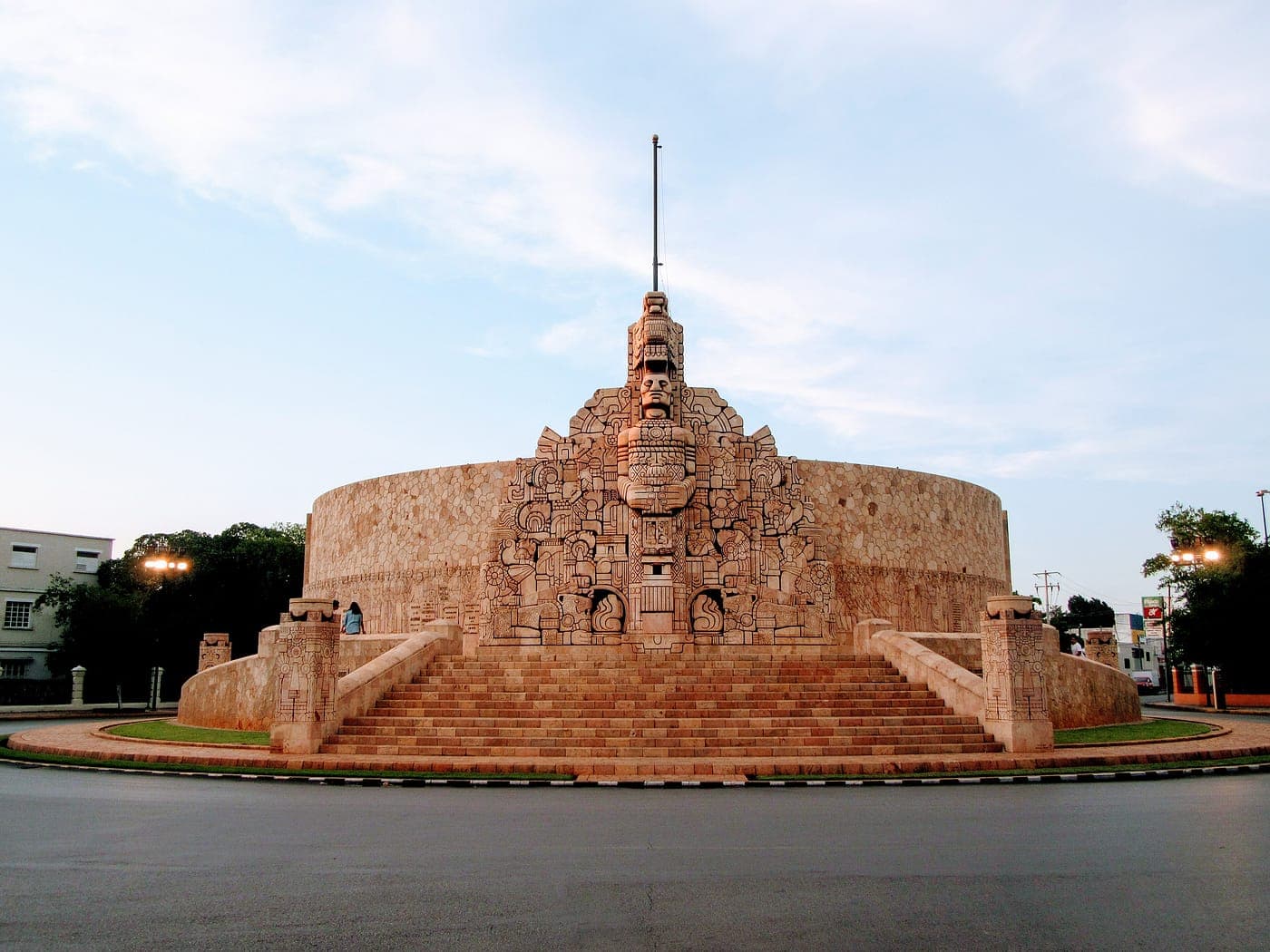 El Monumento a la Patria de Mérida: un muro curvo de piedra tallada con figuras, rodeado de césped y palmeras.