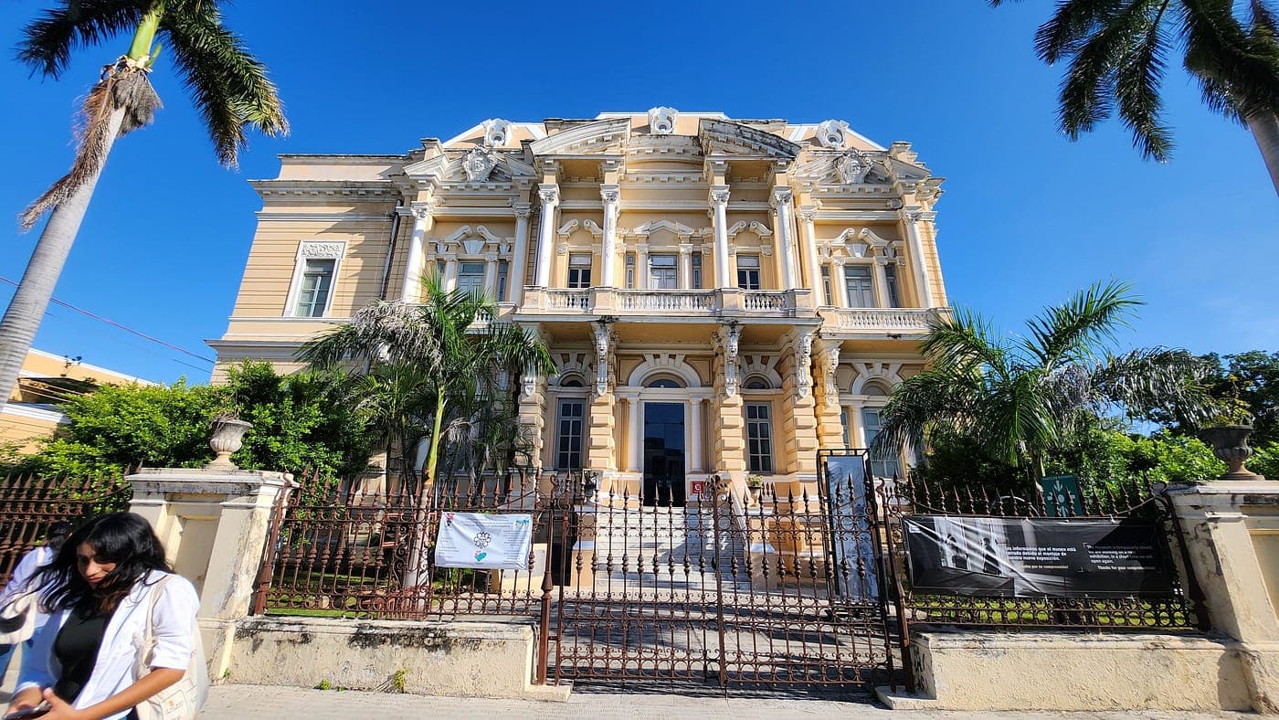 El Palacio Cantón: un palacete afrancesado de piedra clara con columnas y balaustradas, entre palmeras.