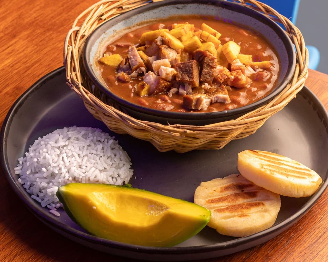 Cazuela de Frijoles: Frijoles cocinados lentamente al estilo paisa, servidos con arroz, aguacate, arepitas, plátano macho y chicharrón crujiente.