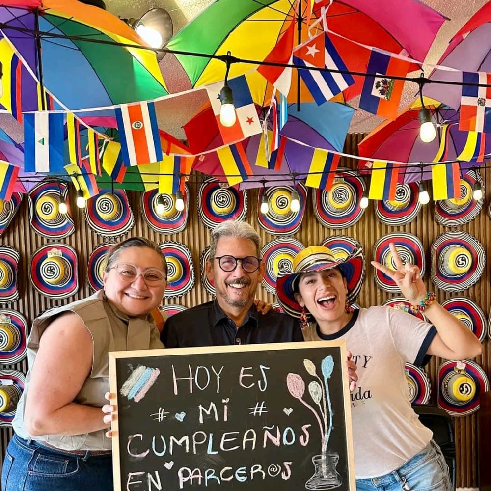 Three people hold up a chalkboard reading «Hoy es mi cumpleaños en Parceros», under the ceiling of colored umbrellas and the wall of sombreros vueltiaos.
