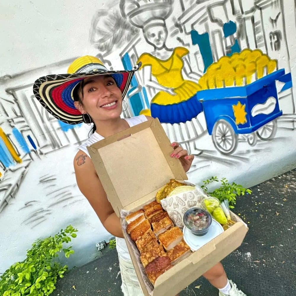 A server in a sombrero vueltiao holds an open box of Castacón — oven-roasted pork, guacamole and salsa — in front of the mango-cart mural.