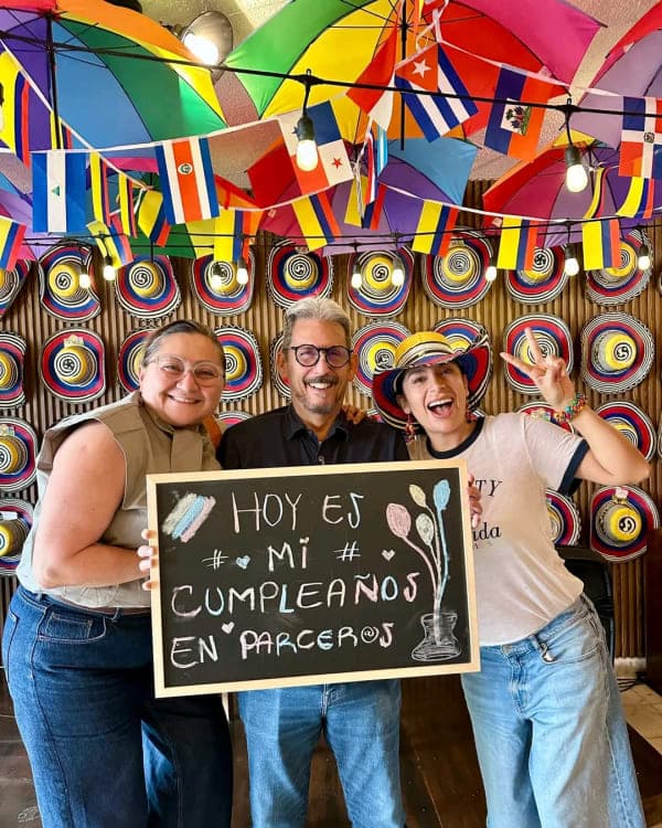 Three people hold up a chalkboard reading «Hoy es mi cumpleaños en Parceros», under the ceiling of colored umbrellas and the wall of sombreros vueltiaos.