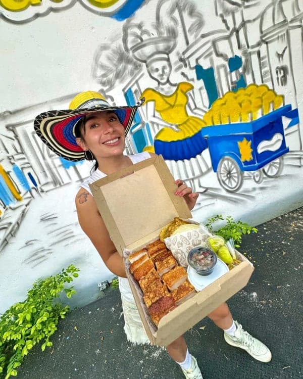 A server in a sombrero vueltiao holds an open box of Castacón — oven-roasted pork, guacamole and salsa — in front of the mango-cart mural.